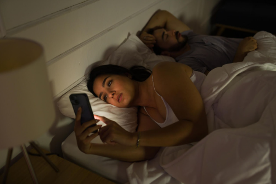 Woman using smartphone in bed at night causing eye strain while partner sleeps